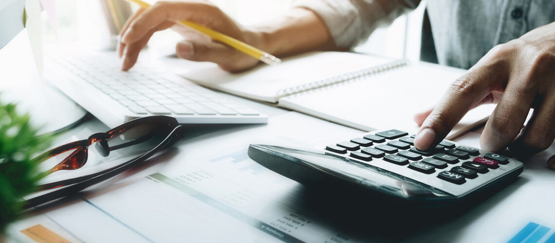 A close-up view of a person working at a desk, with one hand typing on a white keyboard and the other pressing a button on a black calculator. A yellow pencil is held between the fingers. A pair of sunglasses lies on top of documents featuring bar graphs and numerical data. A spiral-bound notebook with blank pages is partially visible in the background, along with a cup holding colorful pens and sticky notes. The scene is brightly lit, suggesting a workspace.