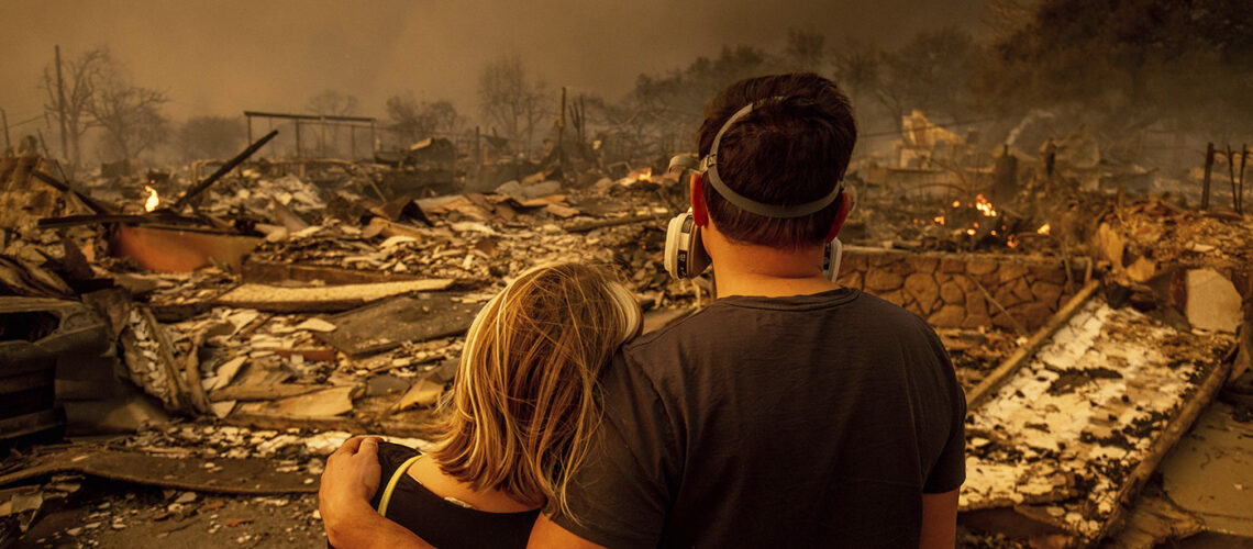 Two people standing, one with their arm around the other, looking at the damage the fires caused.